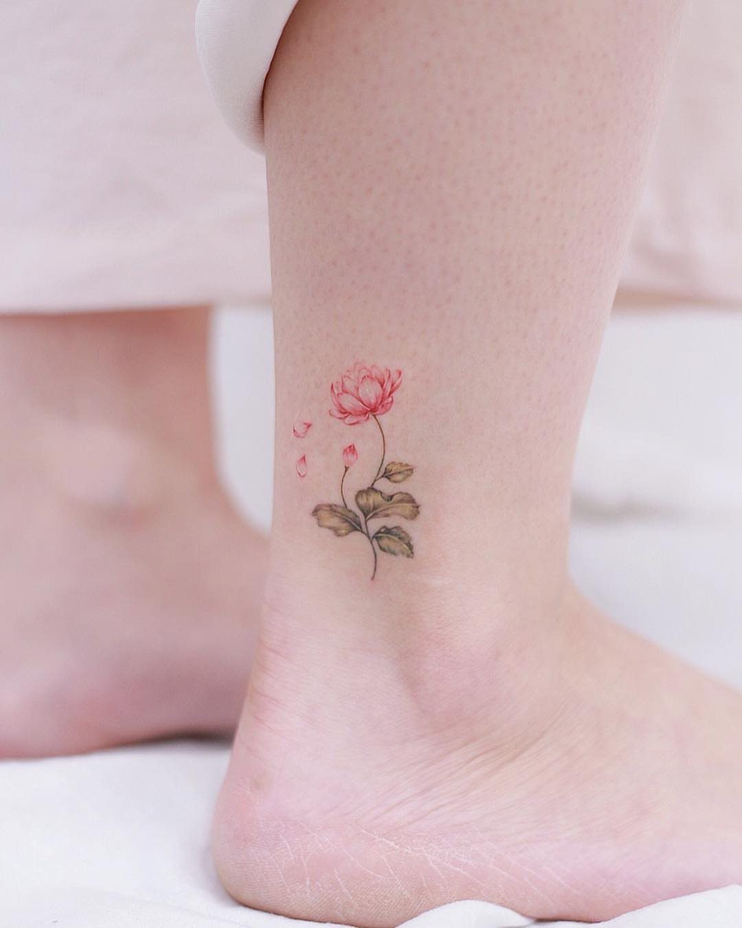 Pink lotus flower tattoo on a person's ankle with green leaves and petals.