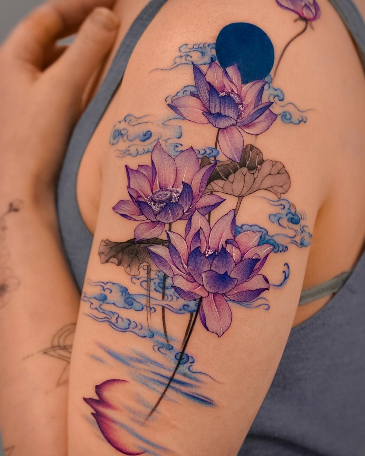 Purple lotus flower tattoo on an upper arm with blue clouds and moon.