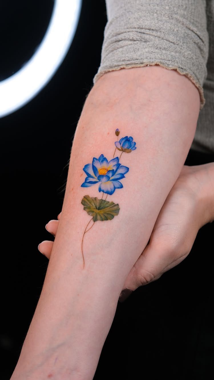 Blue lotus flower tattoo with a green leaf on a person's forearm