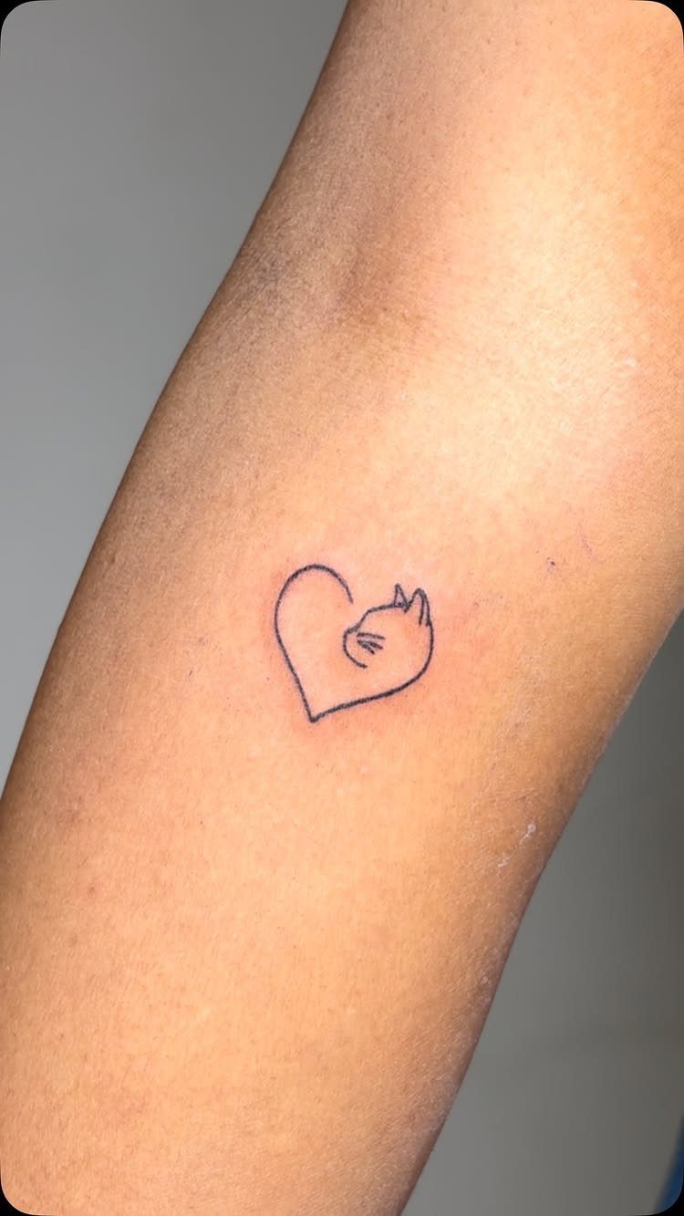 Small heart-shaped cat tattoo with a single line design on a forearm.