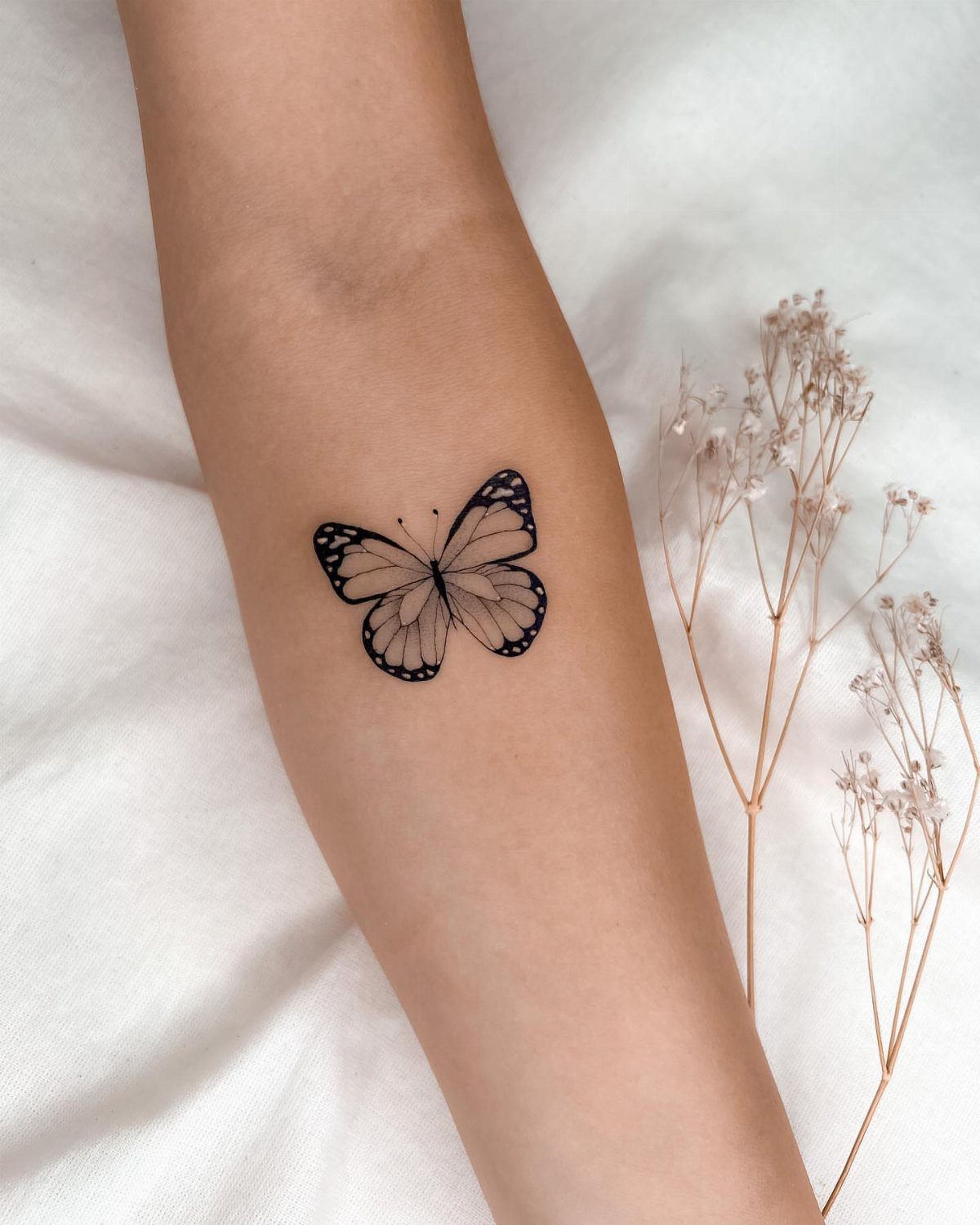 Monarch butterfly tattoo with wings mid-flight on a person's forearm next to dried flowers