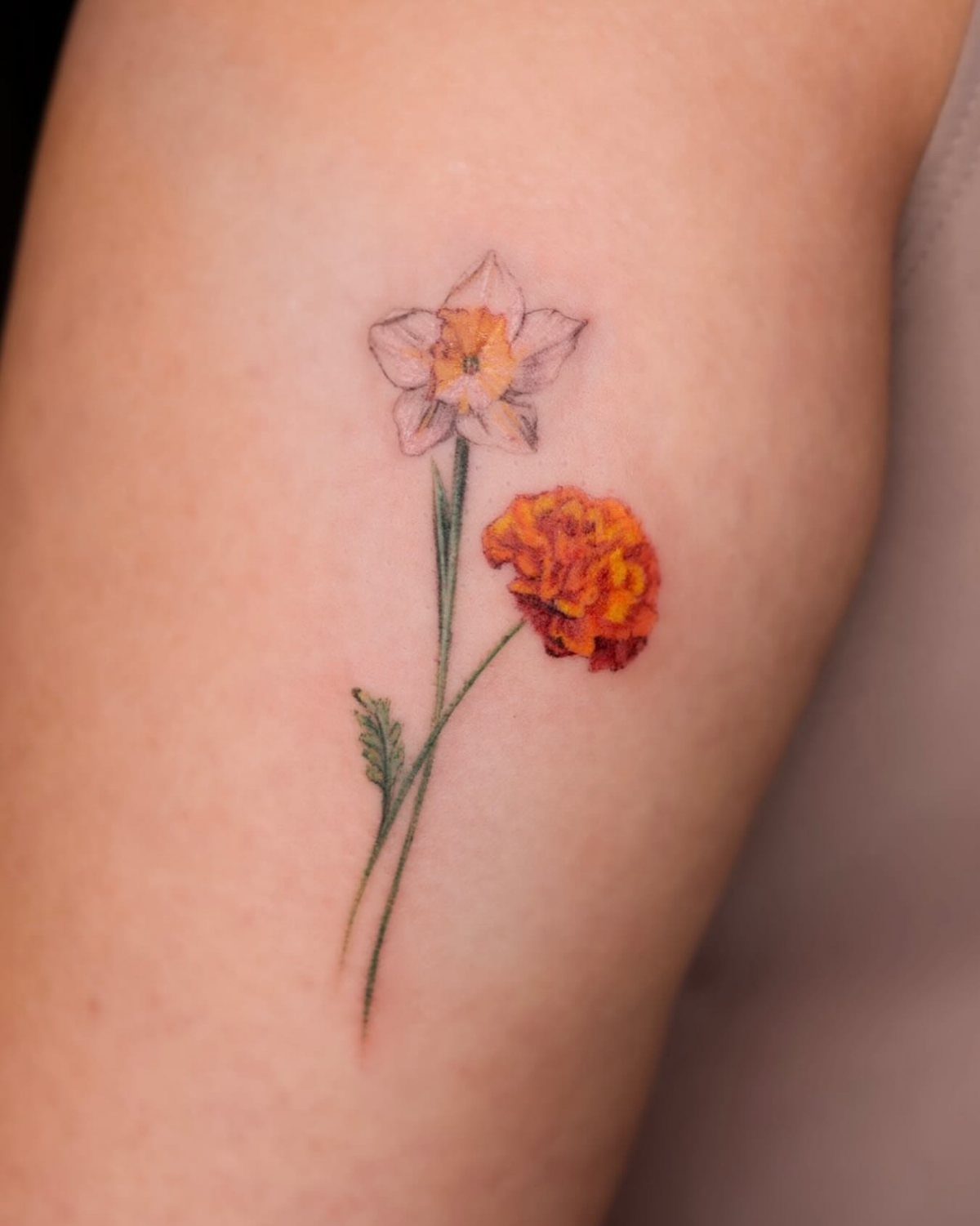 Tattoo of a pale yellow narcissus and orange marigold on a person's arm.