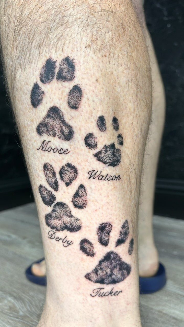 Four black paw print tattoos with names on a person's lower leg