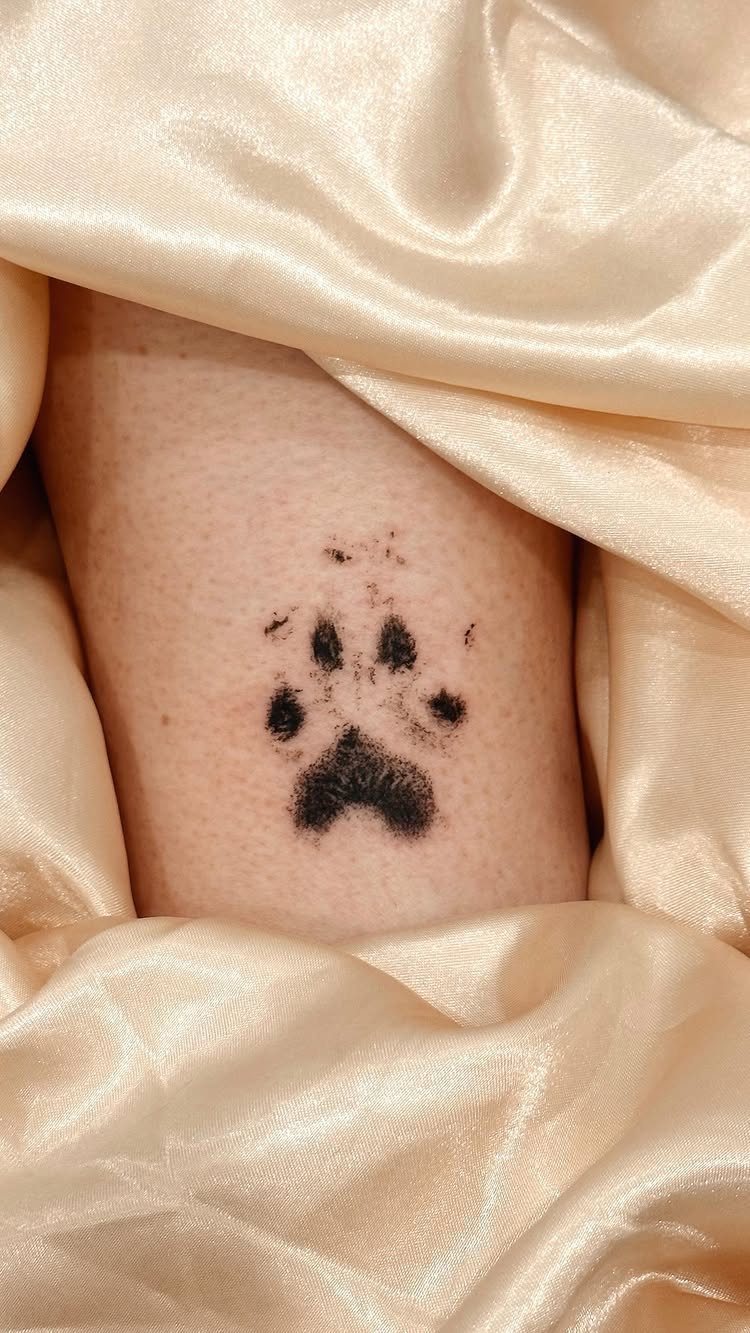 Realistic black ink paw print tattoo on skin surrounded by cream satin fabric.