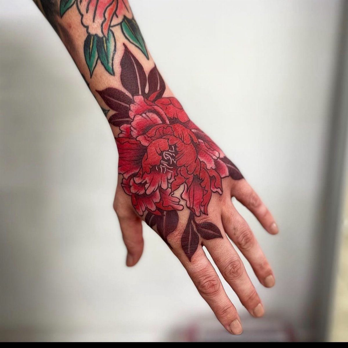 Vibrant red peony flower tattoo with dark leaves on a person's hand.