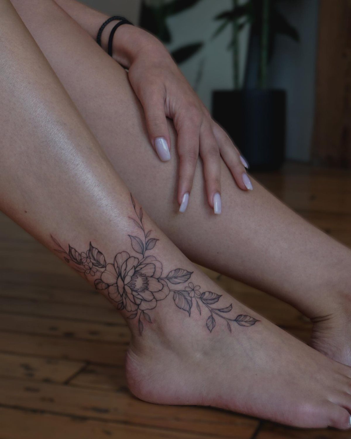 Peony flower linework tattoo wrapped around a person's ankle on a wooden floor.