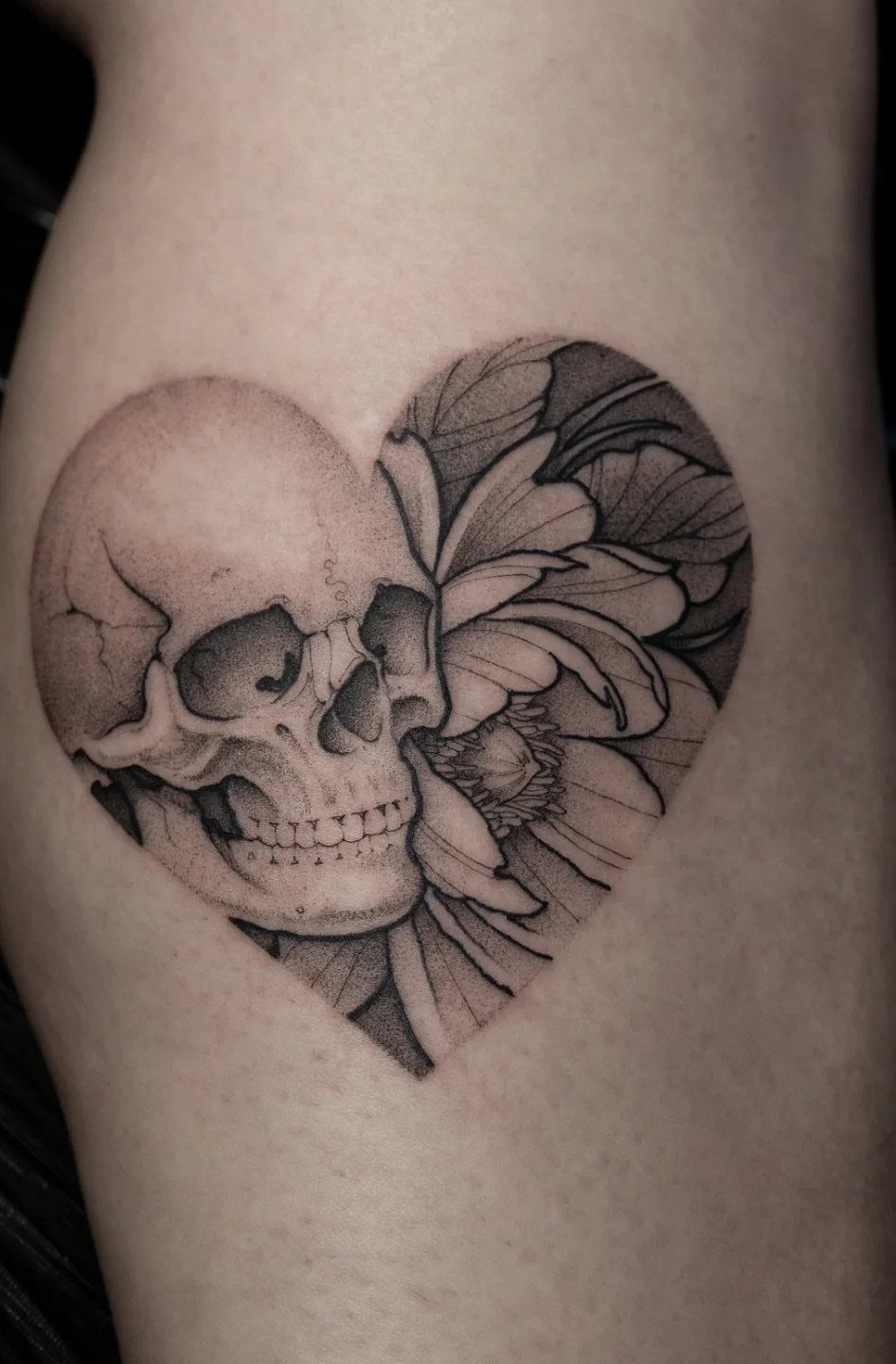 Heart shaped tattoo featuring a human skull and a blooming peony flower.