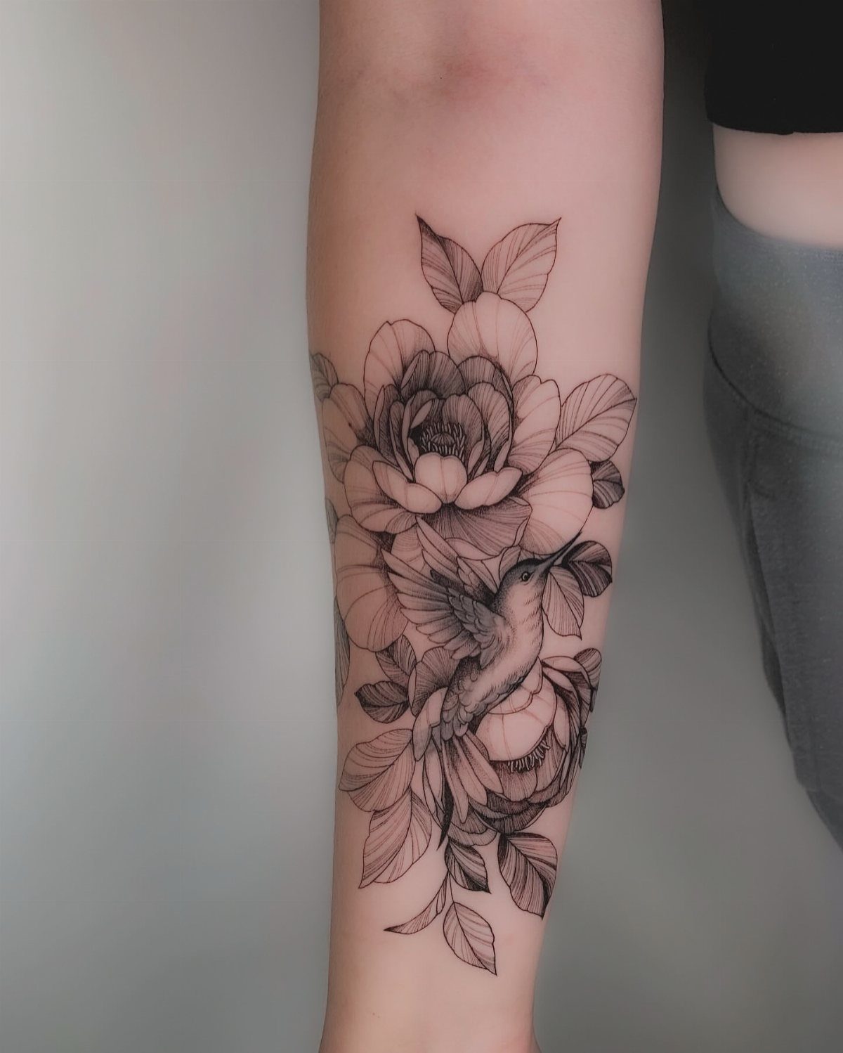 Black and gray tattoo of peony flowers and a hummingbird on a forearm.