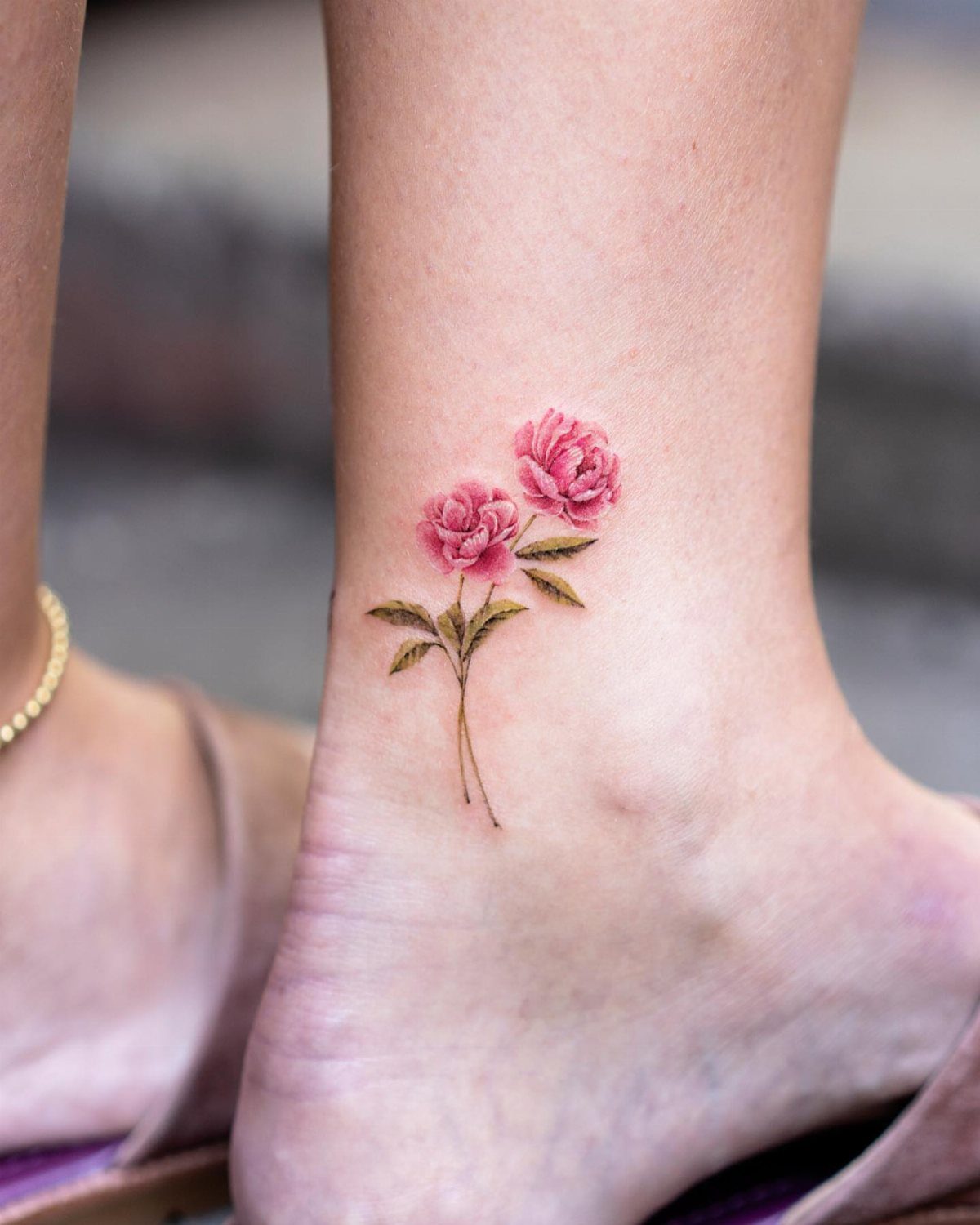 Two pink peony flower tattoos with green leaves on a person's ankle.