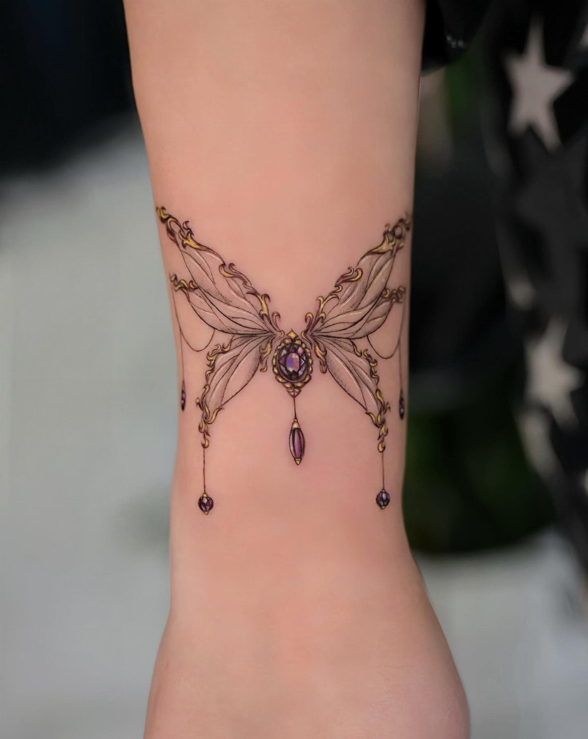 Stylized butterfly tattoo on a wrist with gold filigree and purple gemstones.