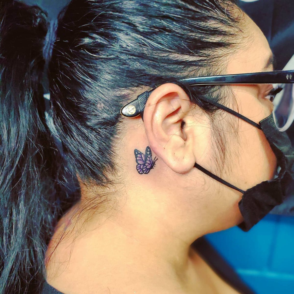 Small purple butterfly tattoo located on the skin behind a person's ear.