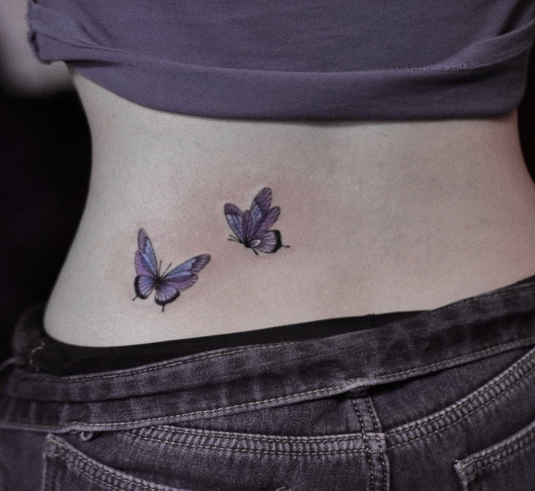 Two purple butterfly tattoos on a person's lower back above grey jeans.