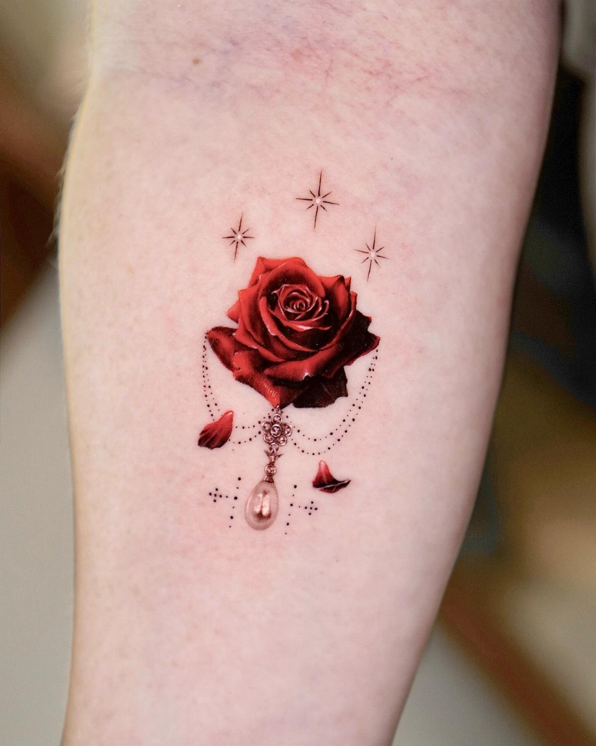 Red rose tattoo with a pearl charm and small stars on a forearm
