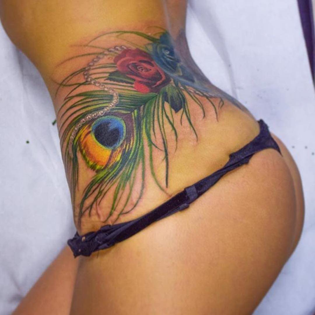 Red rose and peacock feather tattoo on a person's lower back and hip.