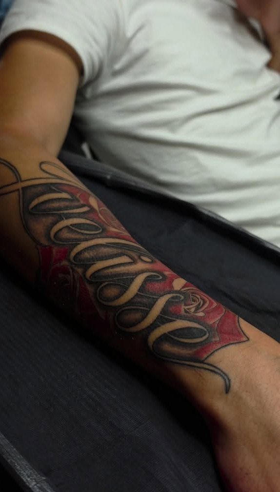 Forearm tattoo featuring a red rose with large cursive text layered on top.