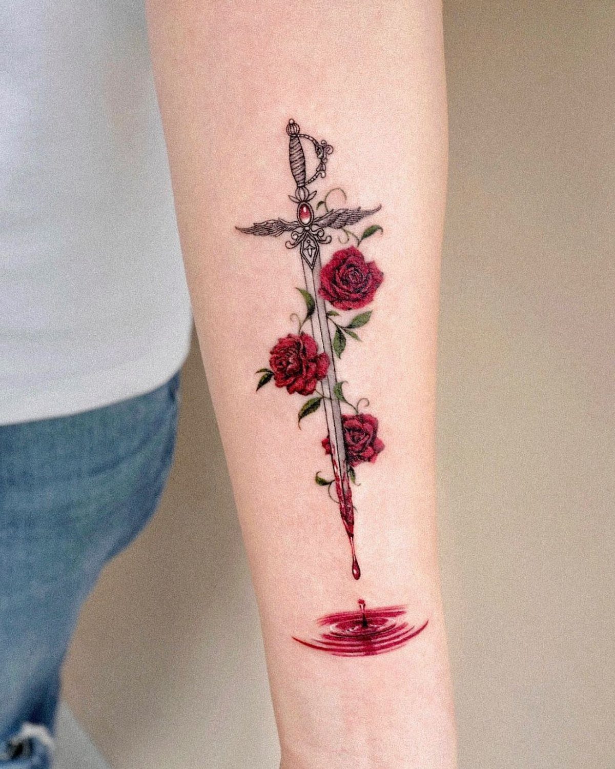 Sword piercing through red roses tattoo on a person's forearm with blood puddle