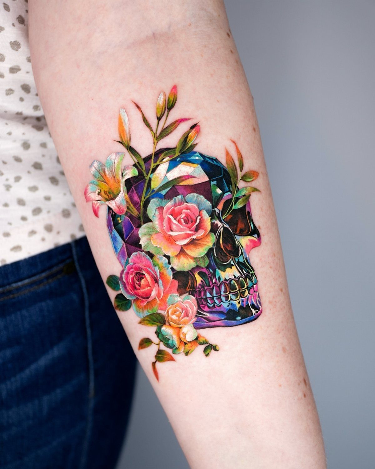 Geometric crystalline skull tattoo with vibrant pink roses and green leaves on forearm