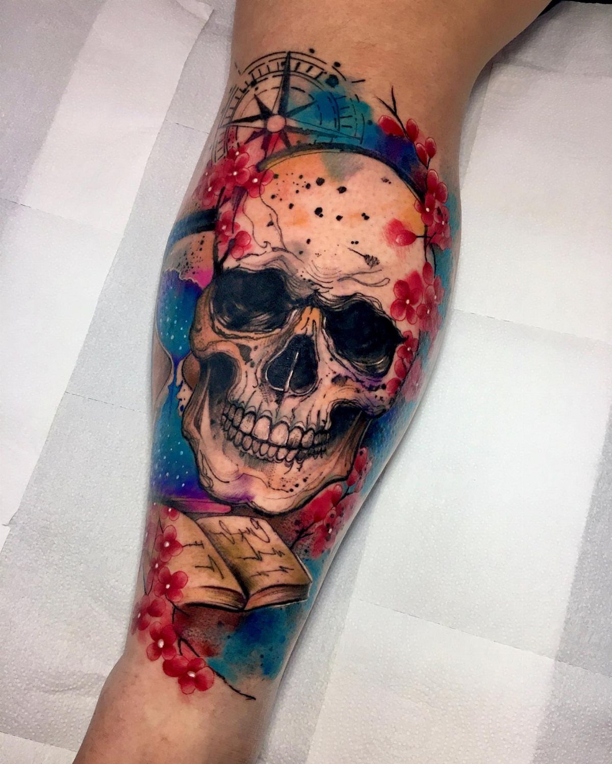 Detailed skull tattoo with red flowers, a compass, and a book in watercolor style.