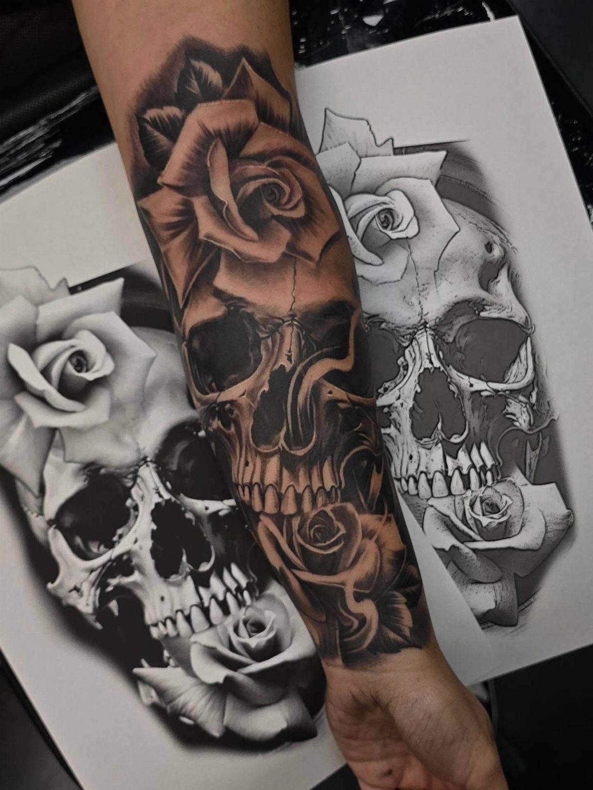 Skull tattoo with a blooming rose on a forearm next to design sketches