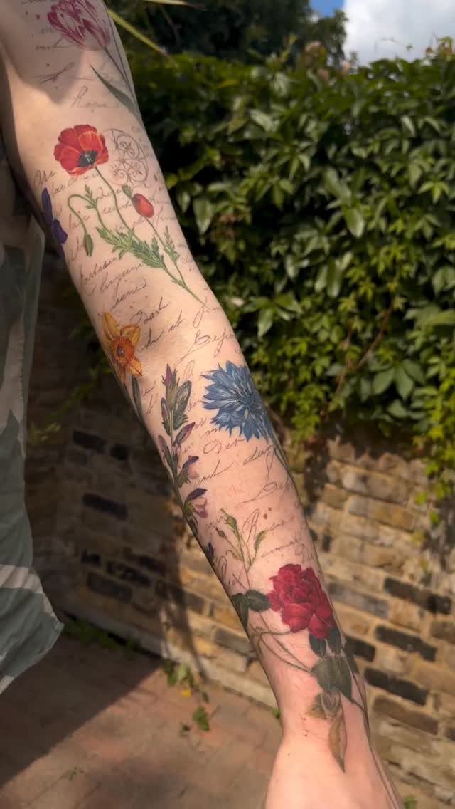 Arm sleeve tattoo featuring red roses, poppies, and blue flowers with cursive script.