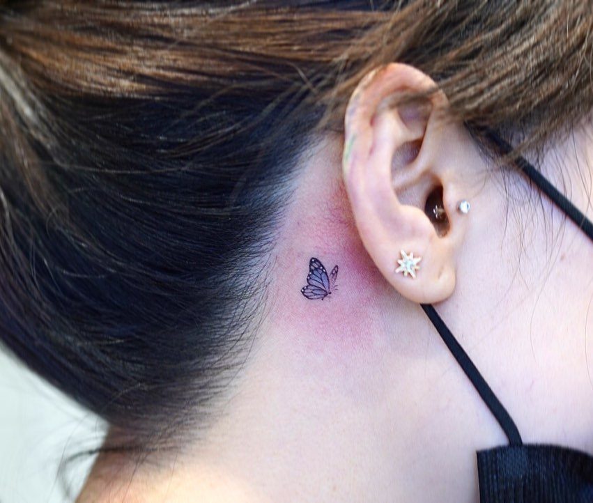 Small blue butterfly tattoo on the neck behind a person's ear.