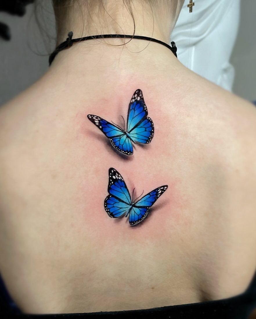 Two 3D realistic blue butterfly tattoos on a person's upper back.