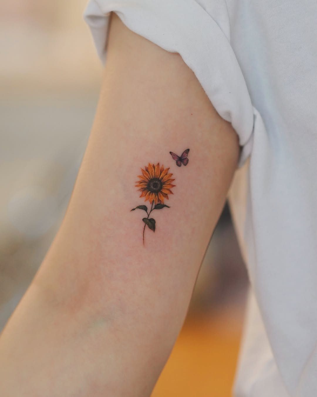 Sunflower and butterfly tattoo on a person's upper arm with vibrant yellow petals.