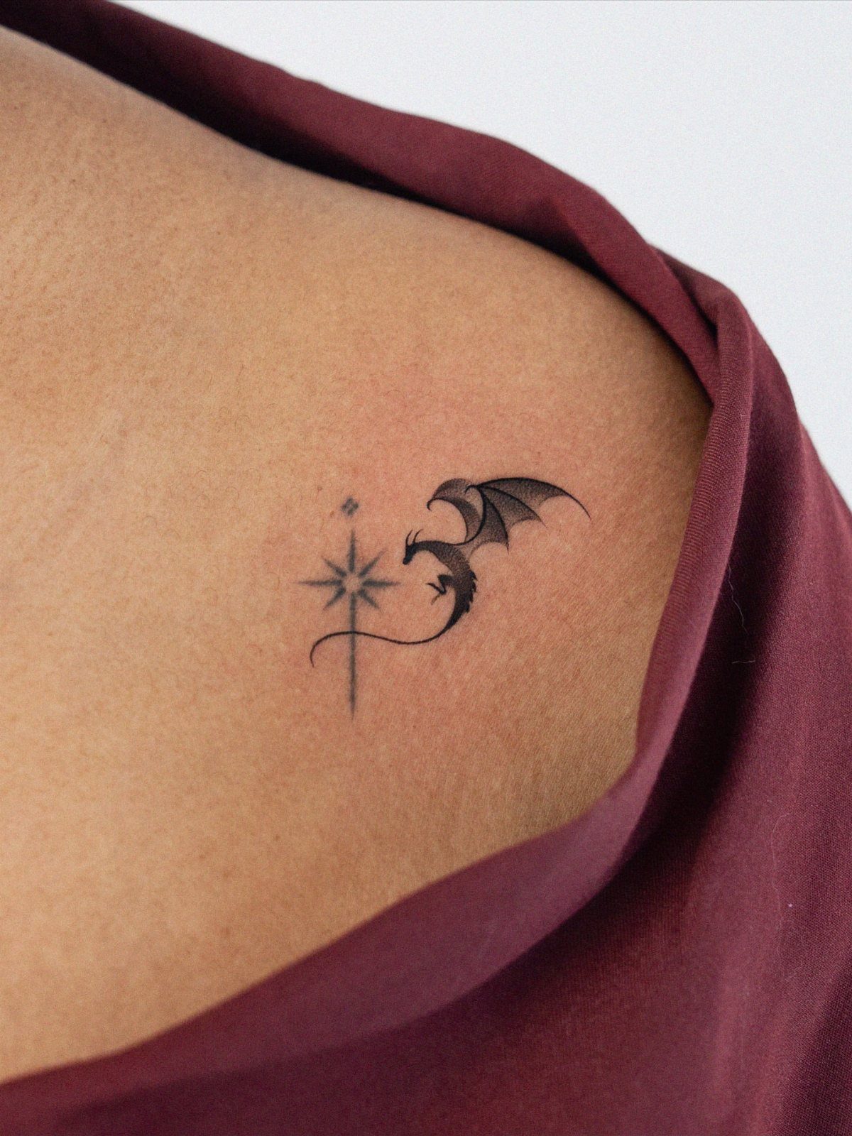 Small black dragon and star tattoo on a person's skin near maroon clothing