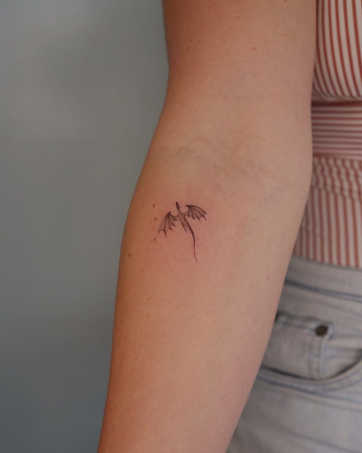 Small black ink dragon tattoo on a person's forearm with outstretched wings.