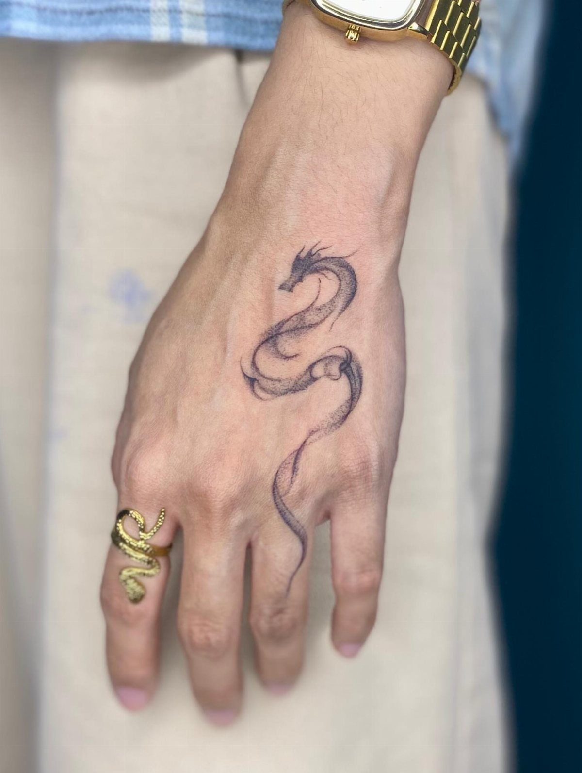 Small black dragon tattoo on the back of a hand with gold jewelry