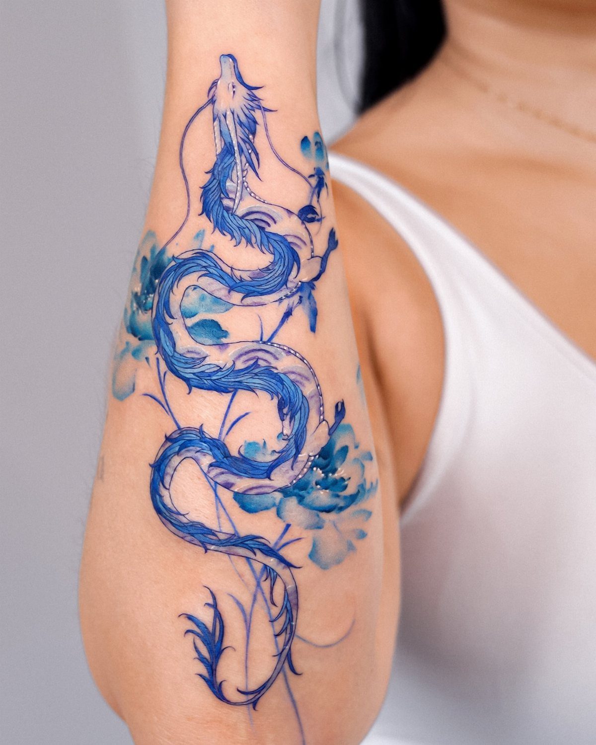 Blue watercolor dragon tattoo with flowers winding down a person's forearm