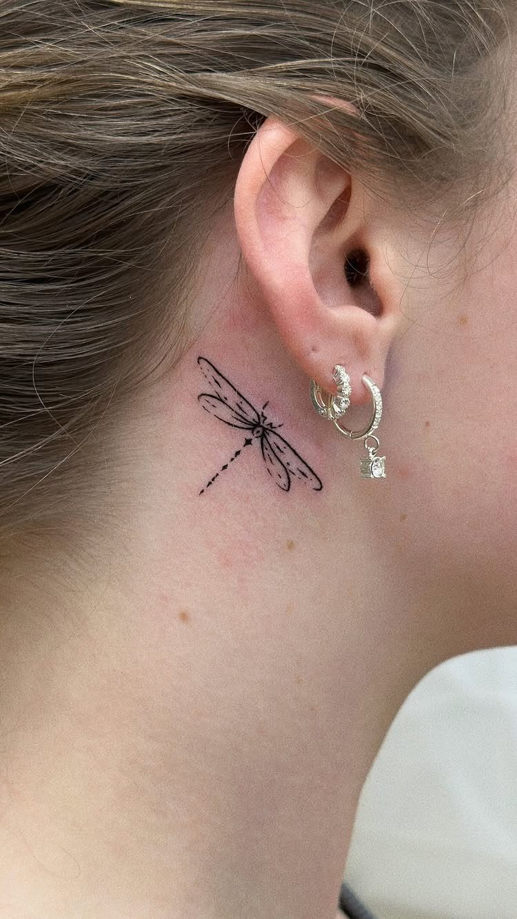 Small black ink dragonfly tattoo on the neck behind a person's ear.