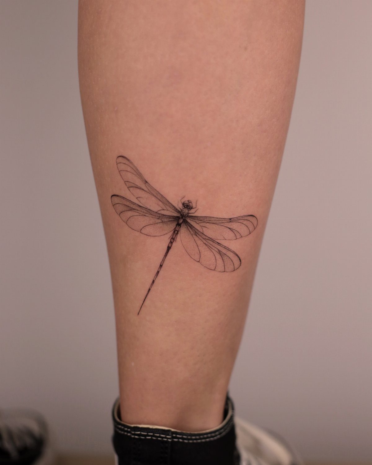Black ink dragonfly outline tattoo on a person's lower leg and ankle.