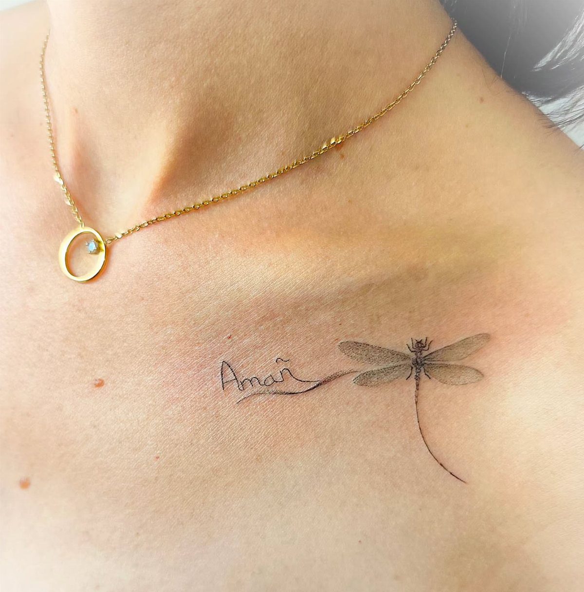 Small dragonfly tattoo with the word Aman on a person's collarbone