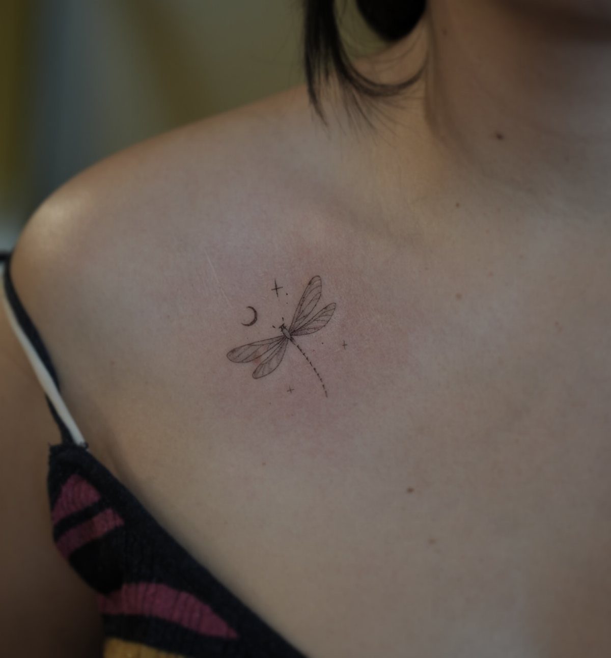 Small dragonfly and crescent moon tattoo on a person's upper chest area.