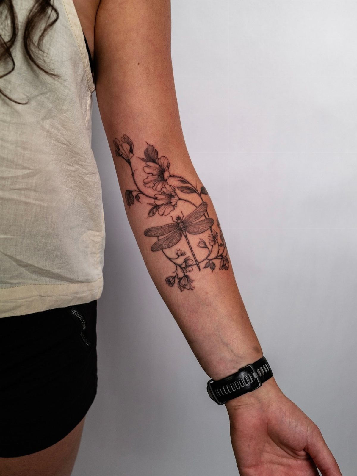 Dragonfly tattoo with flowers and leaves on a person's forearm in black ink.
