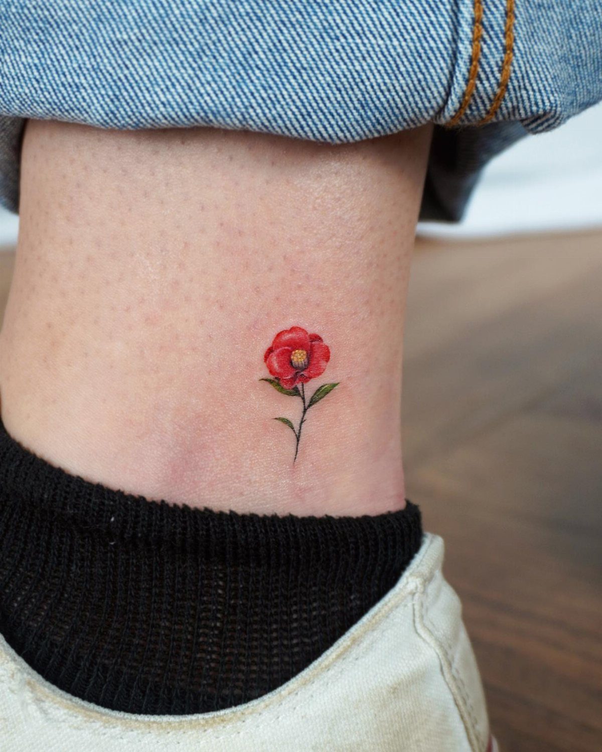 Small red flower tattoo with green stem on a person's ankle