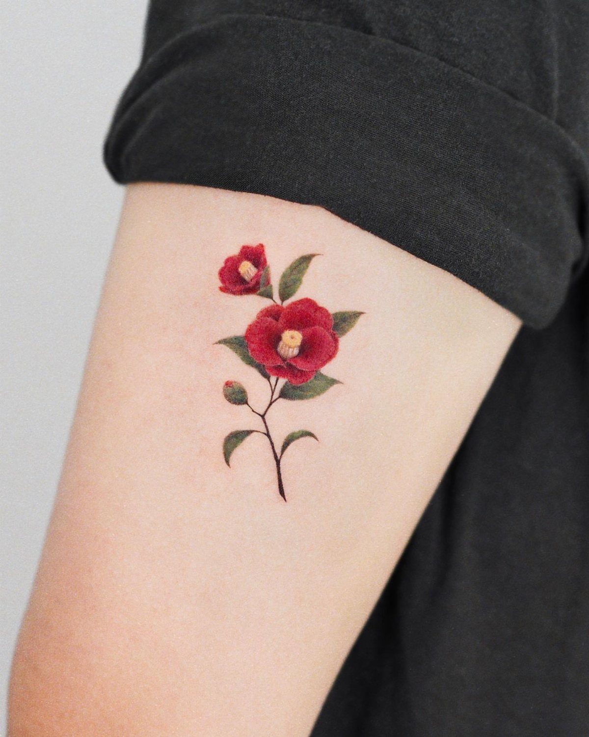 Red camellia flower tattoo with green leaves on a person's upper arm.