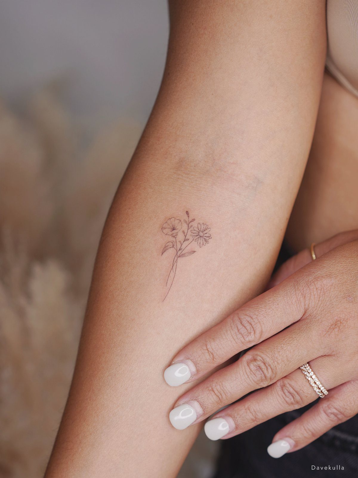 Small wildflower bouquet tattoo on a person's forearm with white manicured nails.