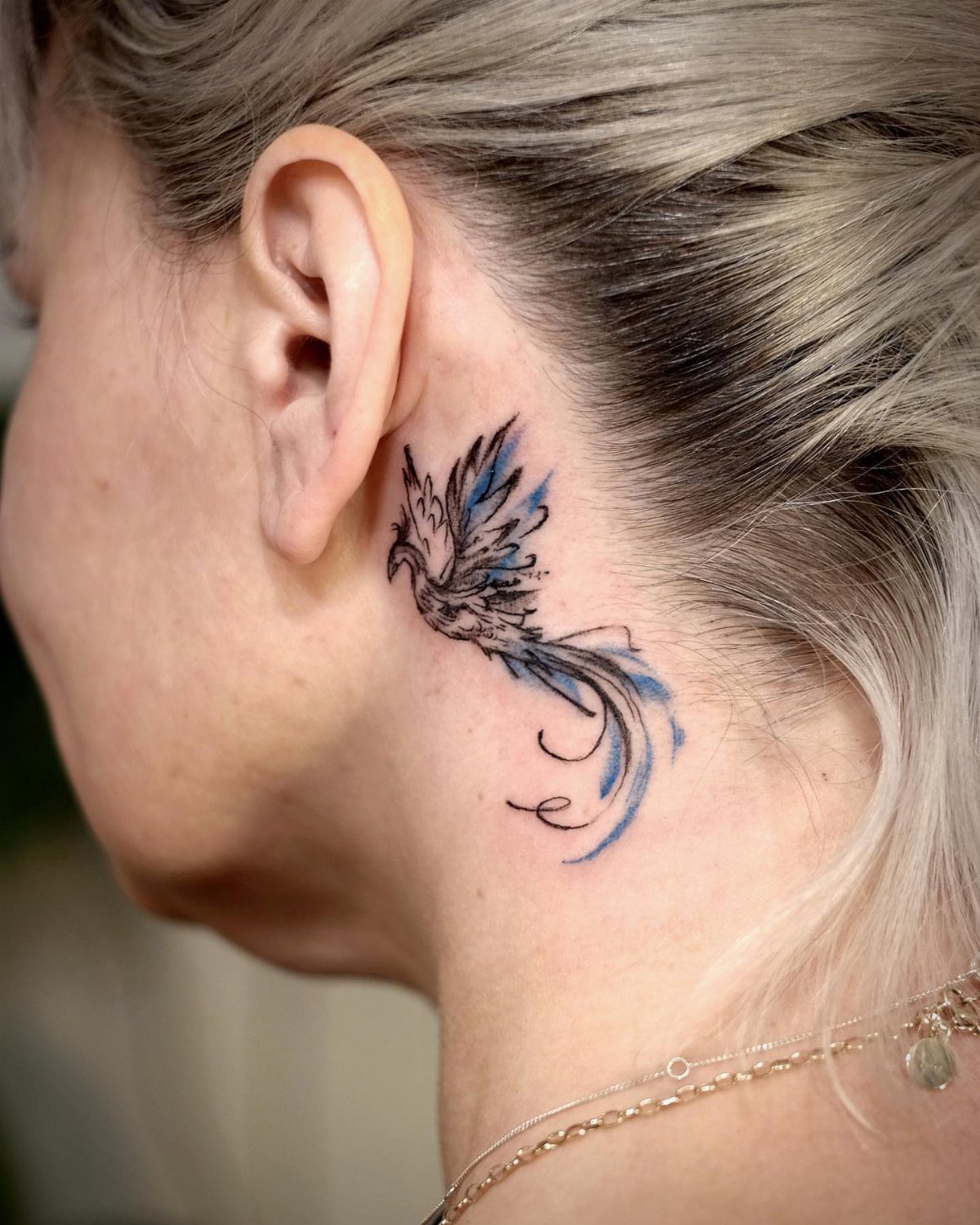 Phoenix tattoo with blue accents placed discreetly behind a person's ear.