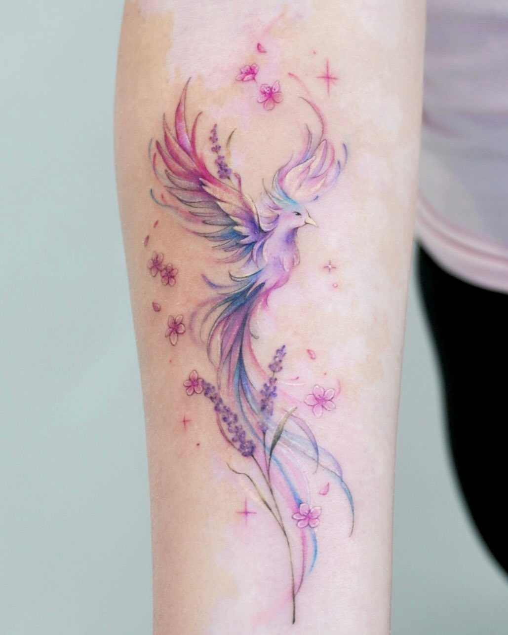 Colorful phoenix tattoo with pink flowers and purple sprigs on a person's arm.