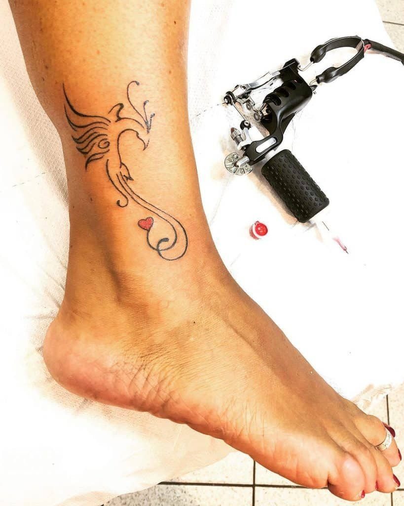 Stylized phoenix tattoo on an ankle featuring a small red heart in the tail
