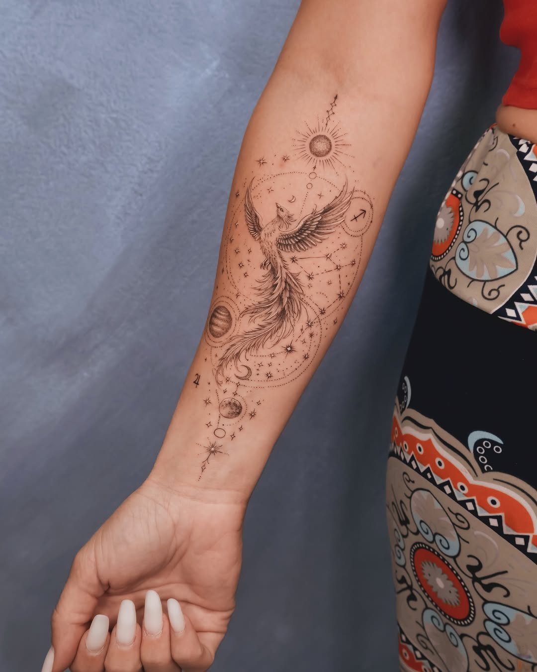 Phoenix tattoo on a forearm surrounded by stars, a sun, and a moon.