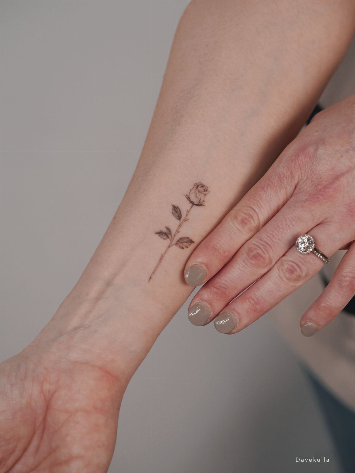 Small fine-line rose bud tattoo on a forearm next to a ringed hand.