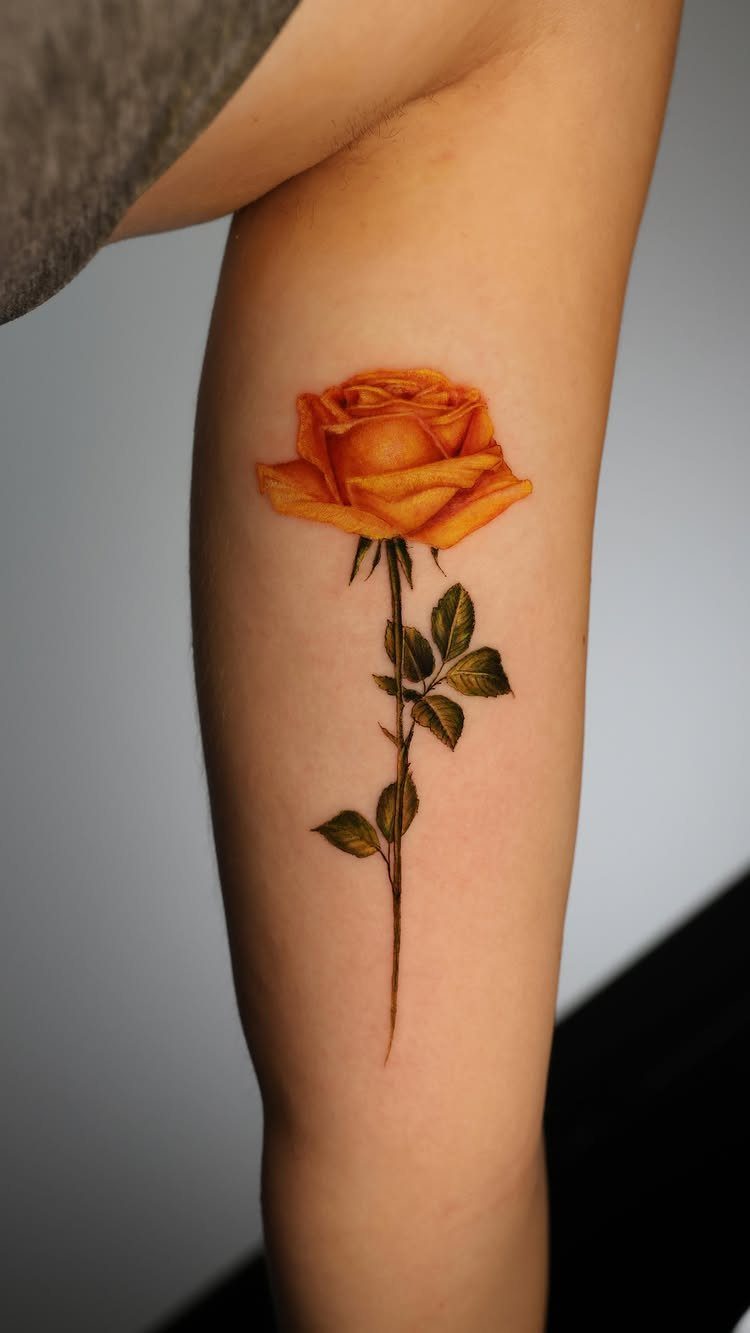 Orange rose tattoo with a green stem and leaves on a forearm