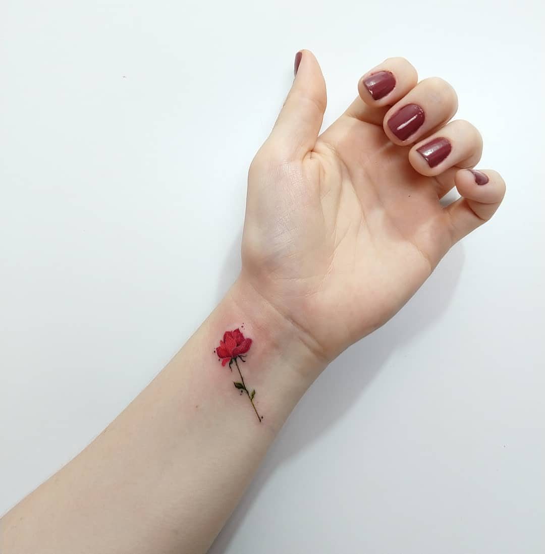 Small red rose tattoo with a green stem on a person's inner wrist