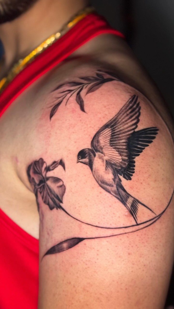 Swallow bird tattoo with floral accents on a person's shoulder in black ink.