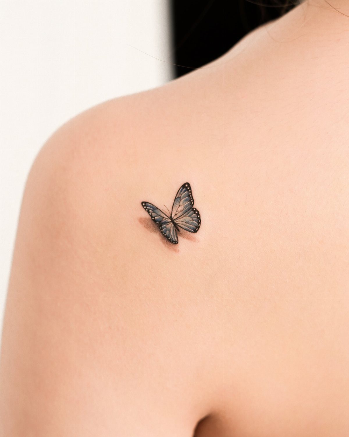 Small butterfly tattoo with soft grey shading on a person's upper back shoulder.