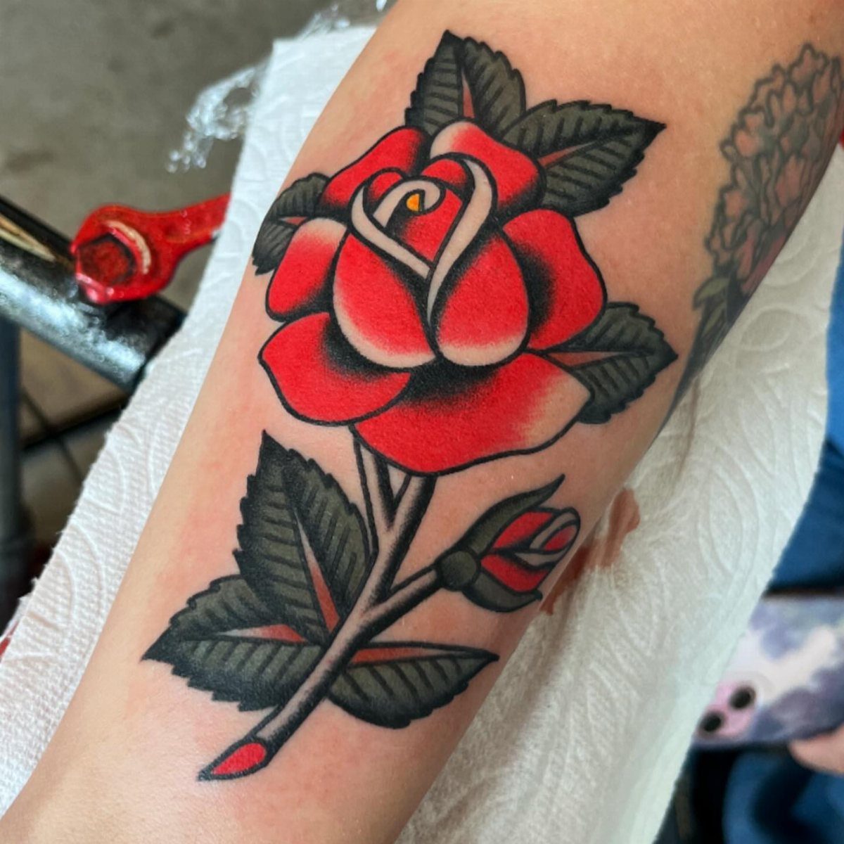 Red rose tattoo with a bud and black leaves on a forearm.