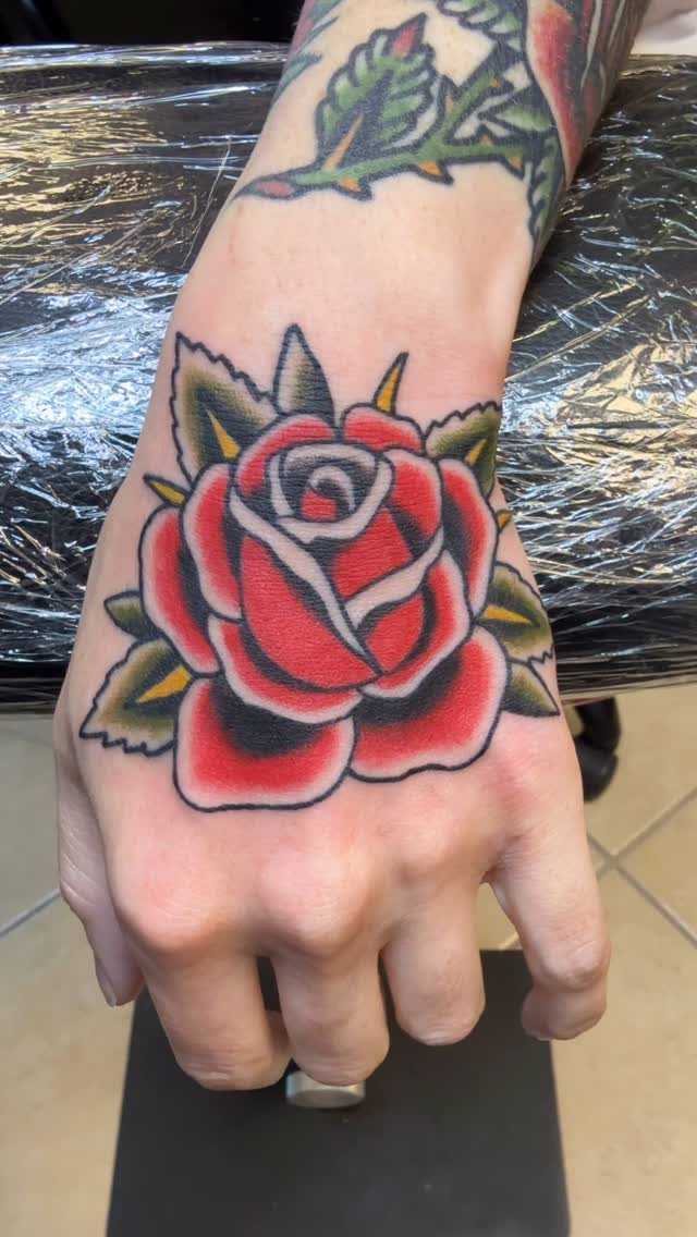 Traditional red rose tattoo with green leaves on the back of a hand.